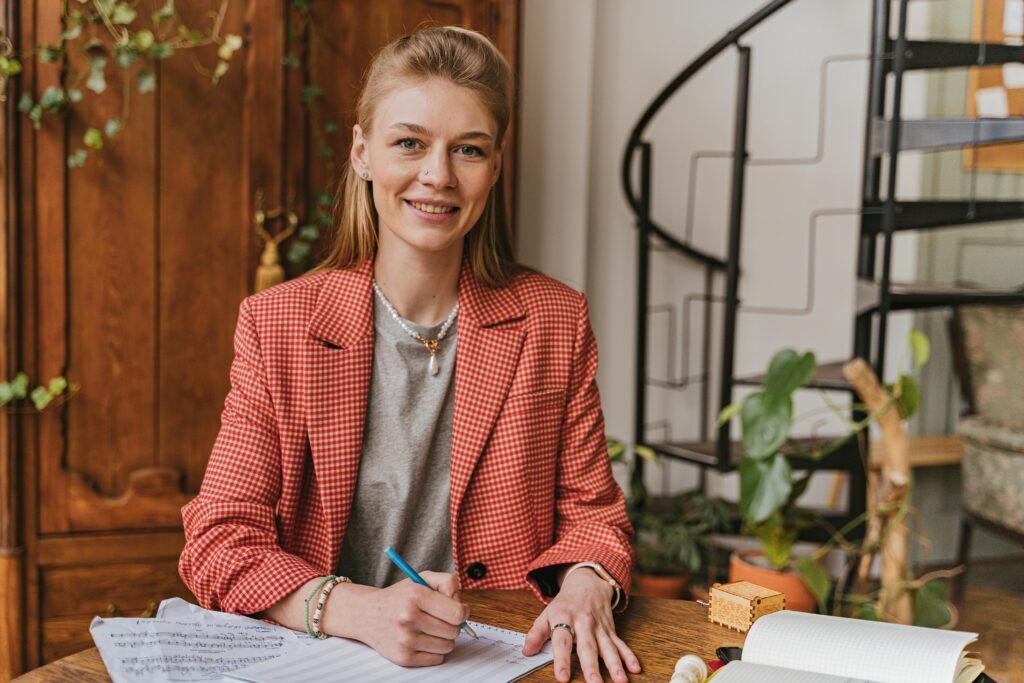 Woman in red blazer smiling while writing at a wooden desk in a cozy home office.
